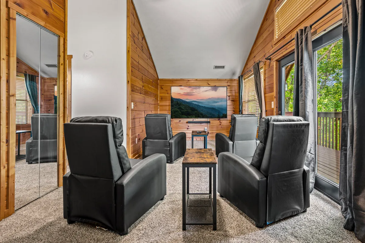 A cozy theater room with plush leather armchairs facing a large wall-mounted screen, a wooden table for snacks or games, and expansive windows with scenic outdoor views. The space exudes comfort, making it ideal for movie nights or intimate gatherings.