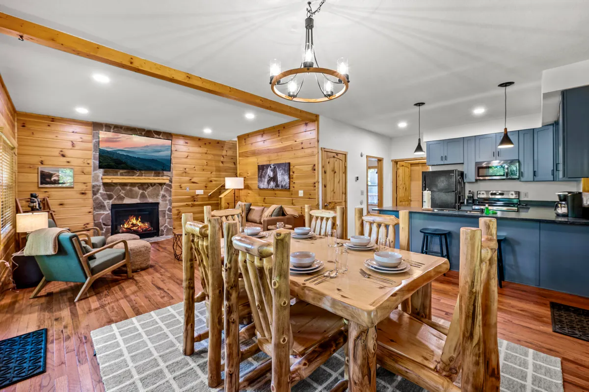 An expansive interior highlights a rustic wooden dining table, a stone fireplace, blue kitchen cabinetry, and a comfortable seating area, all within view. The space is thoughtfully arranged for group activities, shared meals, and relaxing by the fire, with a welcoming, communal feel.