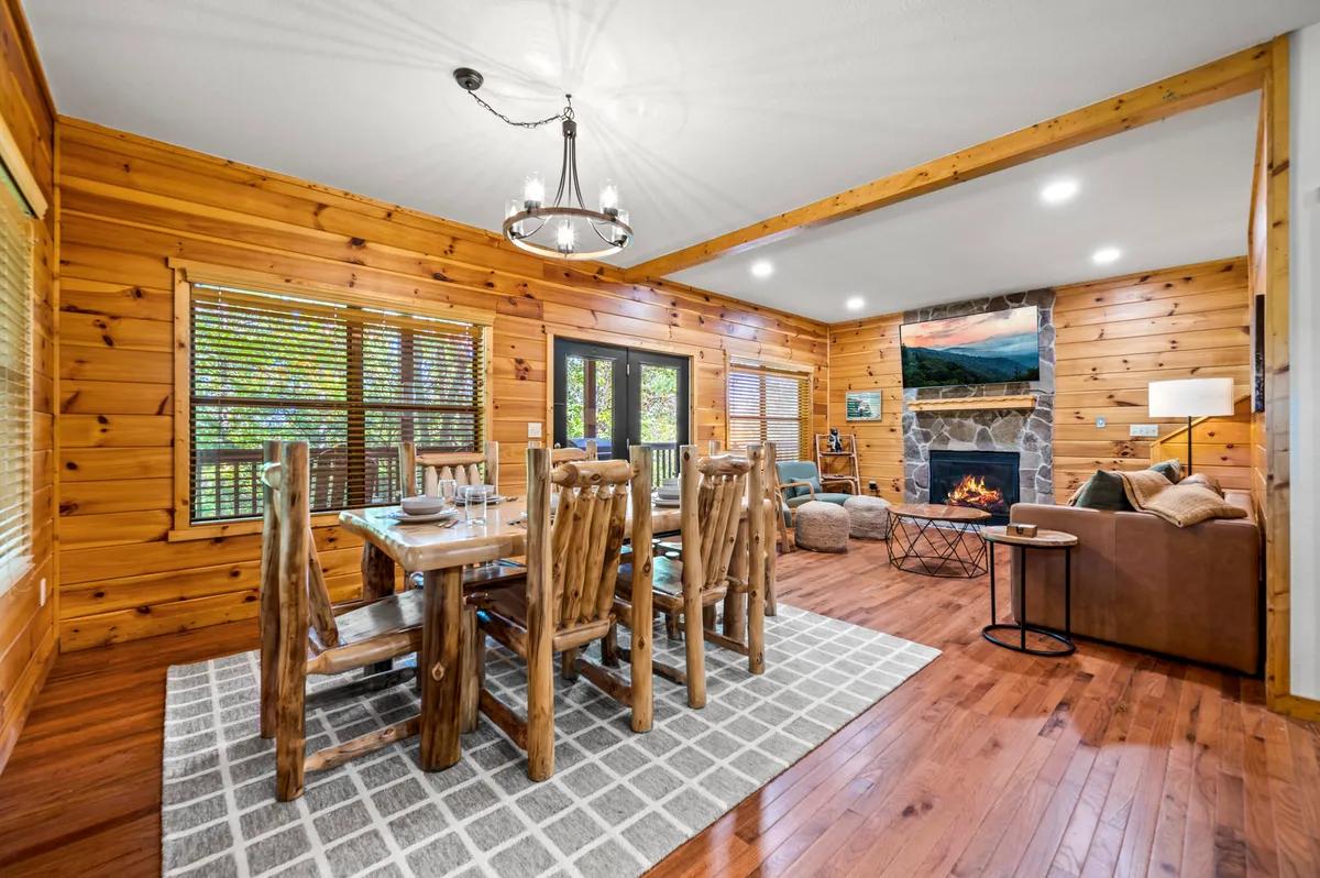 This image showcases a bright, open living and dining space featuring a rustic dining table with chairs, a cozy seating area with a couch, a stone fireplace, and large windows that flood the space with natural light. The open layout seamlessly connects the living and dining areas, evoking warmth and togetherness.