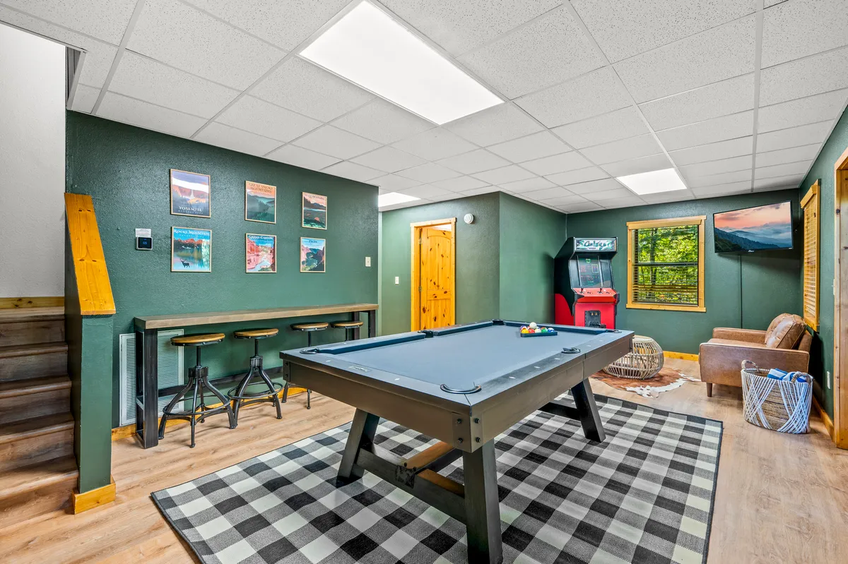 A vibrant game room featuring a classic pool table, bar stools for spectators, a nostalgic arcade machine, framed pictures on the wall, and a window letting in natural light. The setup invites both spirited gameplay and relaxed conversation, appealing to both kids and adults.