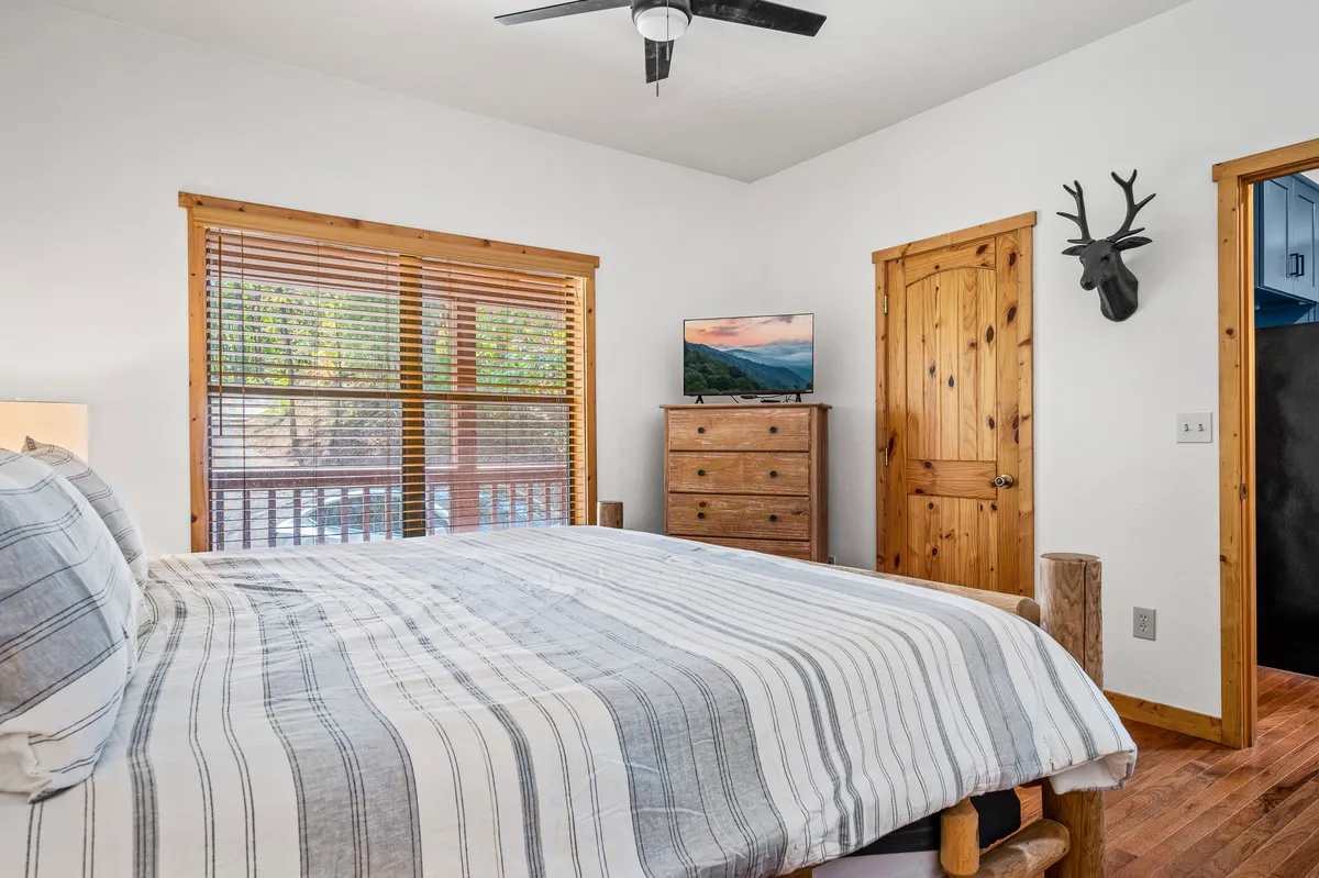 A cozy bedroom featuring a bed with striped bedding, a large window with blinds letting in natural light, wooden furniture, and a television mounted on the wall. Antler wall decor adds a rustic touch, and the window provides a serene view of nature.