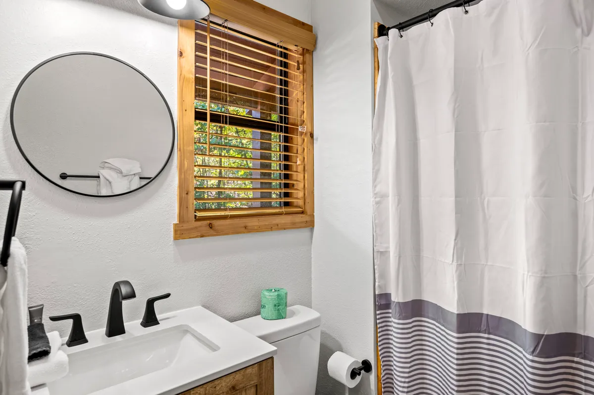 A modern bathroom with a round mirror, a sink with a sleek faucet, wooden accents, and a window with blinds. The space is illuminated by natural light, creating a refreshing and serene environment for personal routines.