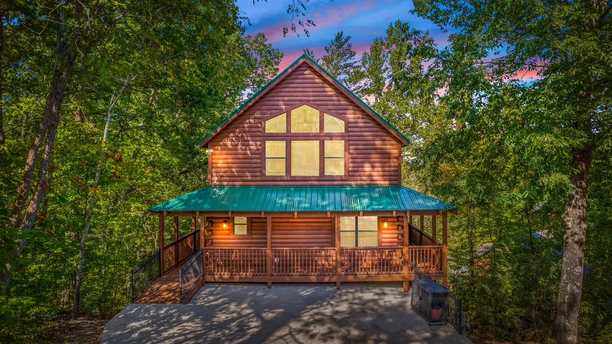 The image showcases the inviting exterior of a classic log cabin nestled in the woods, featuring a green metal roof, expansive wooden porch, and large windows glowing with warm light. The porch overlooks a scenic natural backdrop, perfect for sunset views and outdoor relaxation.
