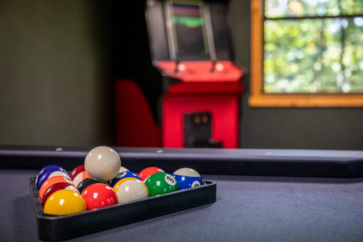 A game room scene where a pool table stands ready for play, with a rack of balls set up, and a classic arcade machine positioned nearby. The lighting and decor create a casual, fun-filled ambiance perfect for tournaments and group activities.
