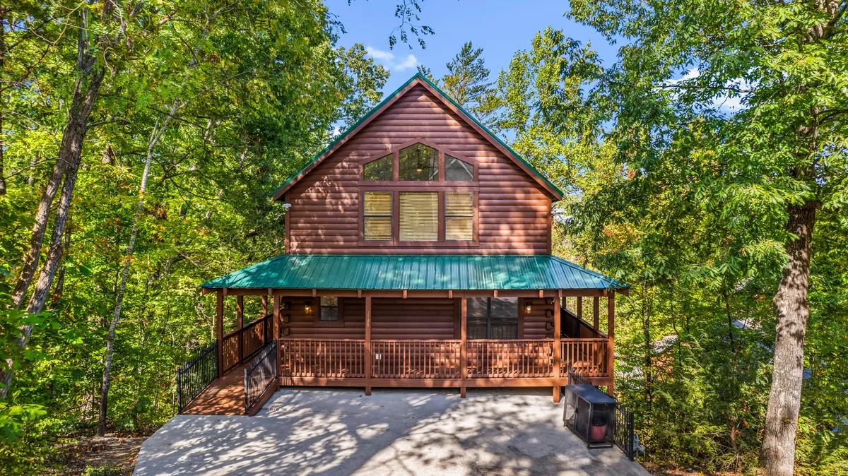 This image features a classic wooden cabin structure nestled among tall trees, with a green metal roof and a large front porch perfect for gathering. The porch is spacious, ideal for family relaxation after a day of adventure, and the surrounding forest hints at the beautiful, natural environment just outside the cabin.