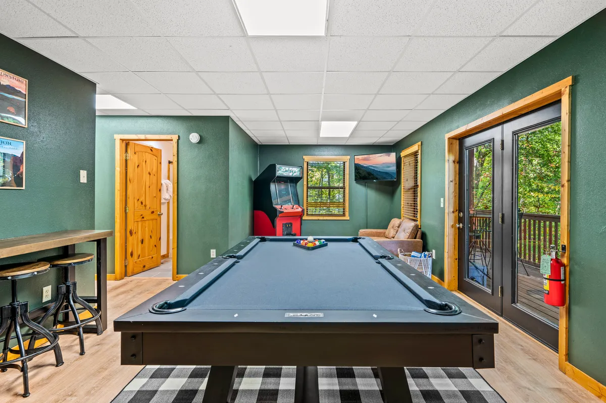 An inviting game room setup with a pool table, an arcade machine, a plush sofa, and windows letting in natural light. The space looks both fun and comfortable, ideal for family game competitions or winding down together indoors after an active day.