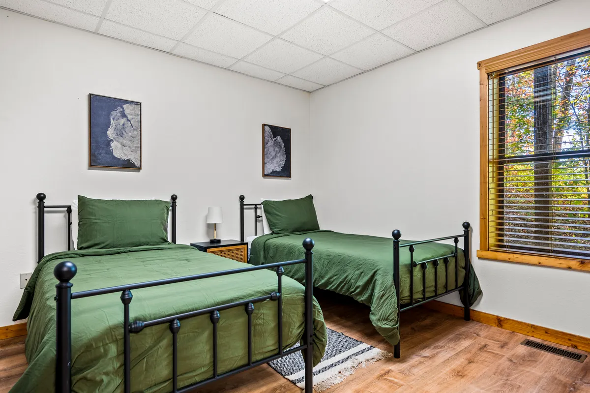 A charming twin bedroom with two beds adorned in green bedding, abstract wall art, and a window featuring wooden blinds. The space is brightened by natural light, offering a relaxing, kid-friendly atmosphere.
