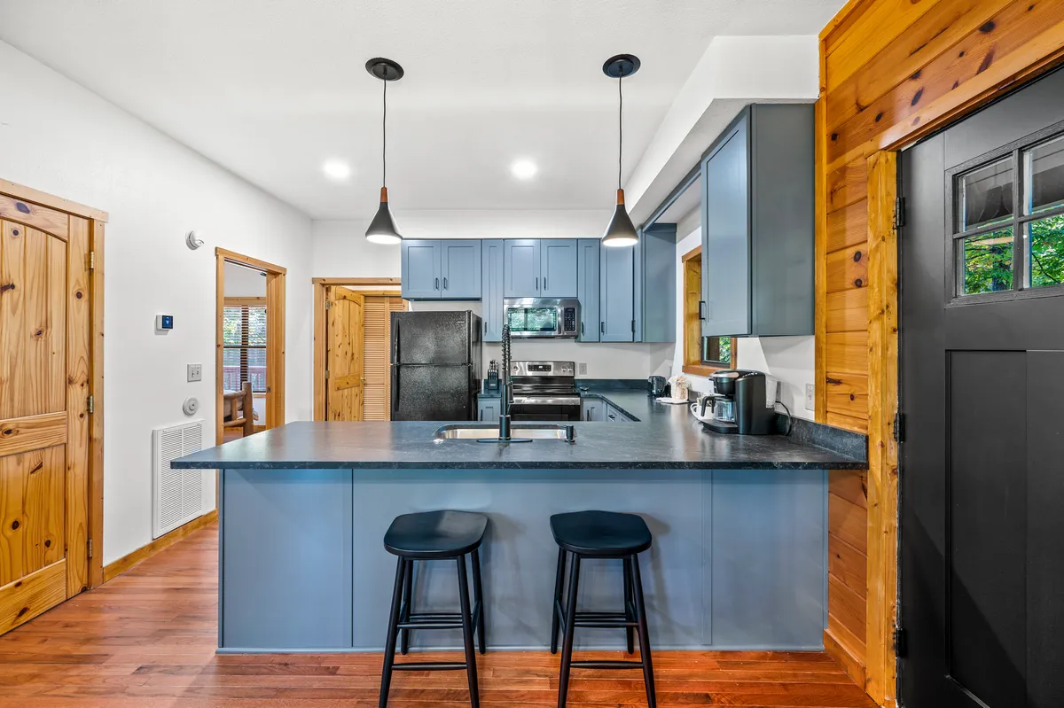 The image features a modern kitchen with a prominent island, blue cabinetry, black appliances, and warm wooden flooring. It’s designed for cooking, entertaining, and casual dining, and offers ample space for multiple chefs or guests to gather.
